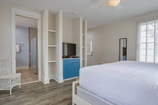 Airy modern bedroom with white bedding, blue media cabinet and flat-screen TV, wicker chair, wood-look floors, large window blinds, and en-suite bathroom with glass shower.