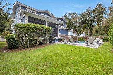 Backyard of a blue two-story house featuring a screened porch, stone patio with a small pool, several lounge chairs and a grill, surrounded by a manicured lawn, trees and a wooden fence.
