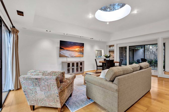 Sunlit open-concept living room with round skylight, hardwood floors, sliding glass doors to a patio, wall-mounted TV showing a beach sunset, patterned armchair and neutral sofa around a rug and dining nook.