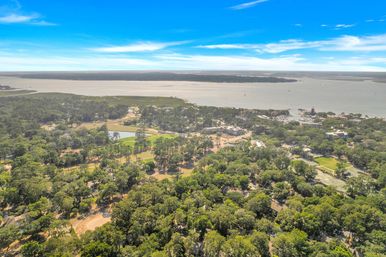 Aerial view of a wooded coastal town and marina on a wide bay with waterfront homes, green parks and ponds under a bright blue sky