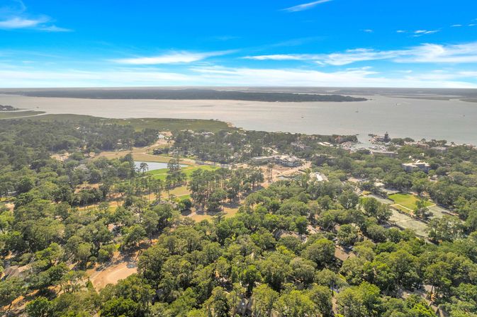 Aerial view of a wooded coastal town and marina on a wide bay with waterfront homes, green parks and ponds under a bright blue sky