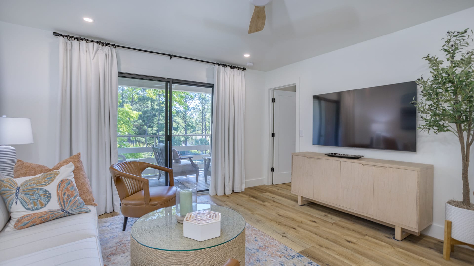 Bright modern living room with wood floors, wall-mounted TV above a light wood console, leather accent chair and round woven coffee table with candle, white sofa with butterfly pillows, sliding glass doors opening to a leafy balcony view