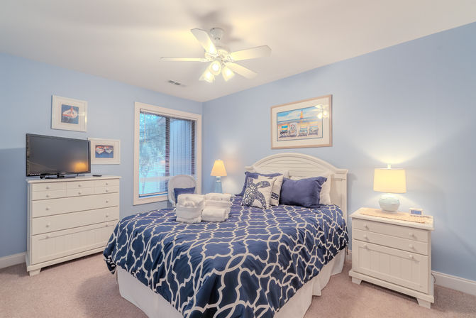 Cozy coastal guest bedroom with light blue walls, white ceiling fan and furniture, navy geometric duvet, folded towels on the bed, beach-themed art, dresser with TV and window blinds.