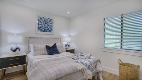 Bright, cozy modern bedroom with neutral plaid bedding and navy accent pillow on a bed, matching mid-century nightstands with glass table lamps, abstract blue wall art, window blinds, woven basket, and hardwood floor.