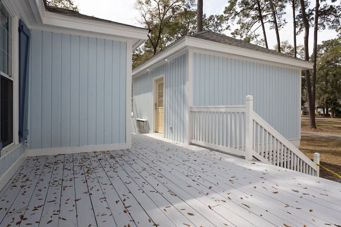 Light-blue coastal-style exterior with white-railed wooden deck strewn with brown leaves, steps to a pale yellow door, and tall pine trees in the yard.
