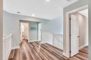 Bright upstairs landing in a modern home with wood-look plank floors, white spindle railing, soft blue-gray walls, and open doors to a bathroom and bedroom.