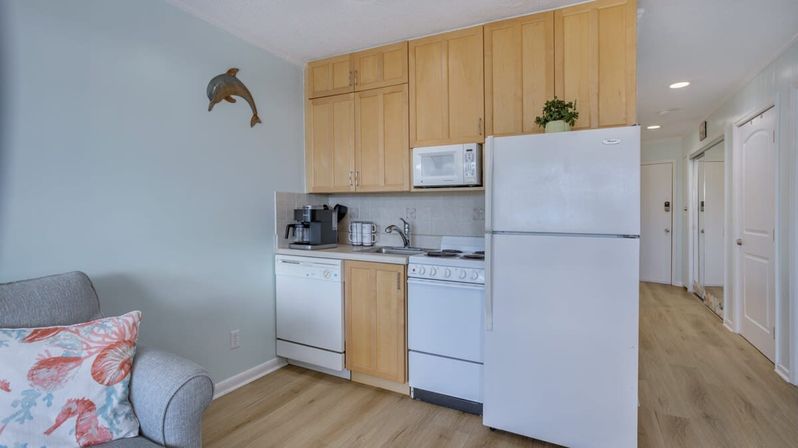 Beach-themed studio kitchenette with light wood cabinets, white fridge and stove, coffee maker, dolphin wall decor, pale blue walls and sofa with seashell pillow