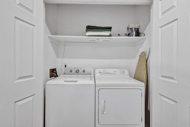 Compact home laundry closet with side-by-side white top-load washer and front-load dryer, white shelf above holding folded green towels and an iron, and a striped ironing board leaning against the wall.