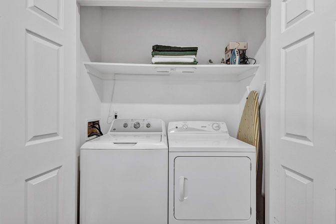 Compact home laundry closet with side-by-side white top-load washer and front-load dryer, white shelf above holding folded green towels and an iron, and a striped ironing board leaning against the wall.