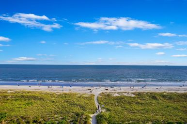 Aerial view of a sunny beach coastline: grassy dunes with a sandy path leading to the shoreline, small waves lapping the sand and scattered beachgoers under a bright blue sky.