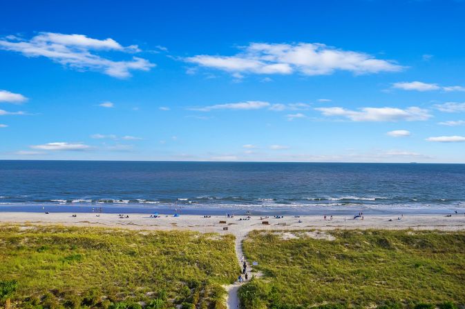 Aerial view of a sunny beach coastline: grassy dunes with a sandy path leading to the shoreline, small waves lapping the sand and scattered beachgoers under a bright blue sky.