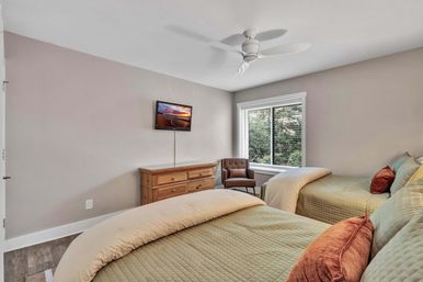 Cozy, sunlit guest bedroom with two twin beds in sage quilted bedding and rust accent pillows, wooden dresser with wall-mounted TV, brown upholstered chair by a large window showing green trees, white ceiling fan and neutral walls.