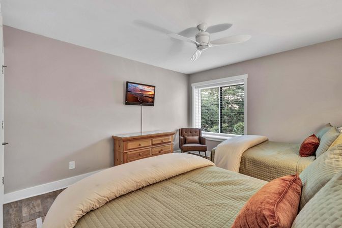 Cozy, sunlit guest bedroom with two twin beds in sage quilted bedding and rust accent pillows, wooden dresser with wall-mounted TV, brown upholstered chair by a large window showing green trees, white ceiling fan and neutral walls.