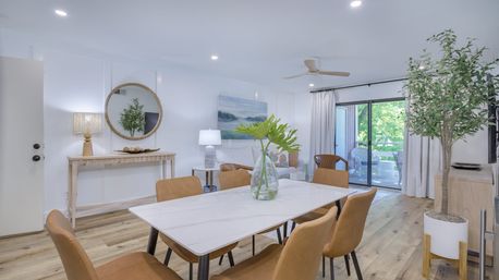 Bright airy open-plan living and dining room with white walls and light wood floors, marble-top dining table with tan chairs and glass vase of green leaves, round mirror over console, potted tree, and sliding glass doors to a leafy balcony.