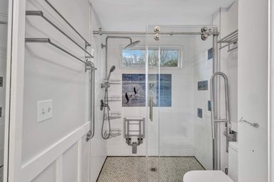 Bright walk-in accessible shower with sliding glass door, stainless grab bars and towel racks, fold-down shower seat, rain and handheld showerheads, white tile walls with blue ocean tile mural, small window and hexagon mosaic floor.