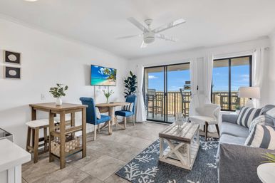Bright coastal condo living room with sliding glass doors to an ocean-view balcony, blue accent chairs, gray striped sofa, rustic wood coffee table, small dining nook, ceiling fan and beach-themed decor