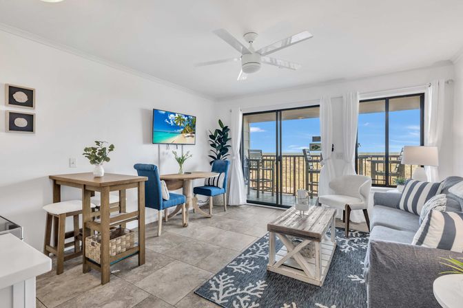 Bright coastal condo living room with sliding glass doors to an ocean-view balcony, blue accent chairs, gray striped sofa, rustic wood coffee table, small dining nook, ceiling fan and beach-themed decor