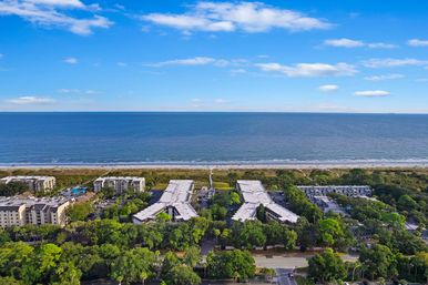 Aerial view of oceanfront condominiums and tree-lined coastal road with sandy beach, calm blue ocean and bright sunny sky
