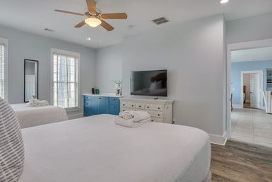 Bright, airy bedroom with two white-made beds topped with folded towels, sunlit windows, blue storage cabinet, wall-mounted TV above a distressed white dresser, wooden ceiling fan and wood-look floors.