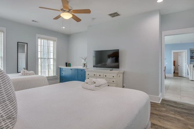 Bright, airy bedroom with two white-made beds topped with folded towels, sunlit windows, blue storage cabinet, wall-mounted TV above a distressed white dresser, wooden ceiling fan and wood-look floors.