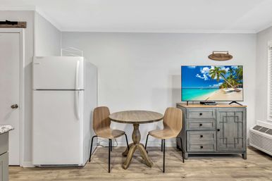 Coastal cottage kitchenette with white refrigerator, round wooden bistro table and two chairs, gray media cabinet with TV showing a tropical beach, and light wood floors — cozy vacation rental vibe.