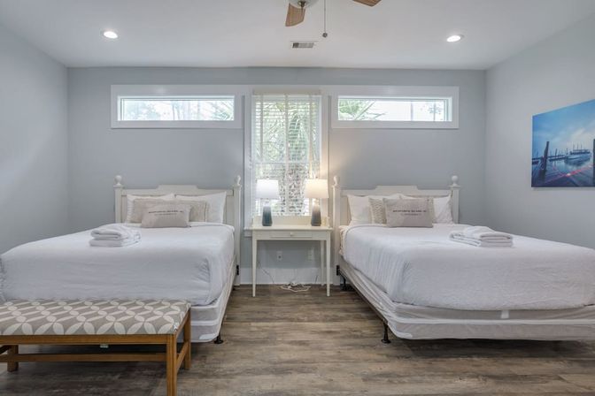 Bright coastal-style guest bedroom with two white beds, central nightstand and lamps, wood-look flooring, transom windows and a palm tree visible outside