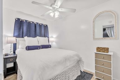 Bright cozy bedroom with white walls and ceiling fan, queen bed in white linens with navy bolster pillows and navy curtains, bedside lamps on a dark nightstand, arched wall mirror and small dresser with wicker basket.