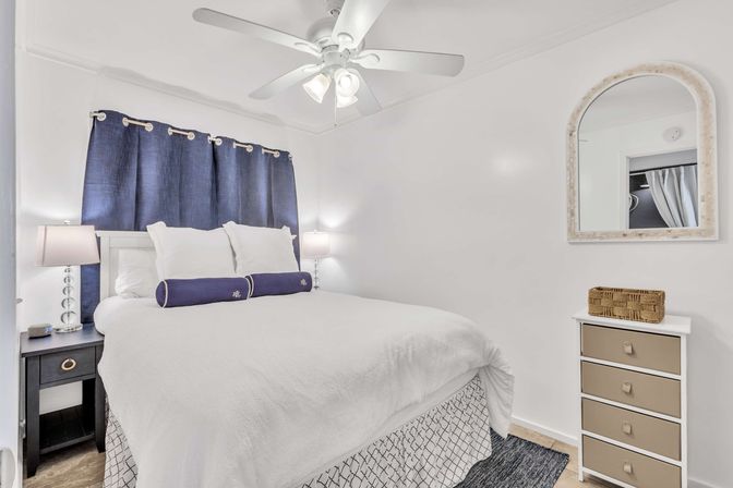 Bright cozy bedroom with white walls and ceiling fan, queen bed in white linens with navy bolster pillows and navy curtains, bedside lamps on a dark nightstand, arched wall mirror and small dresser with wicker basket.