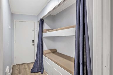 Space-saving built-in bunk beds in a narrow hallway alcove with beige cushions, white paneled walls, navy privacy curtains and light wood flooring