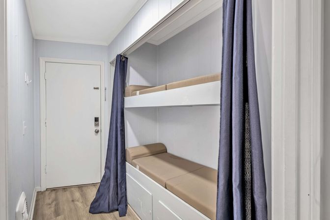 Space-saving built-in bunk beds in a narrow hallway alcove with beige cushions, white paneled walls, navy privacy curtains and light wood flooring