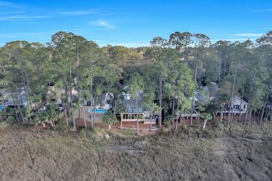 Aerial view of marshfront homes nestled among tall pine trees, featuring a backyard swimming pool, wooden decks and a grassy tidal marsh in the foreground.