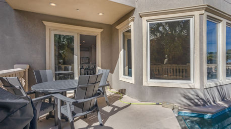 Sunny poolside patio with round grey outdoor table and Adirondack-style chairs on a concrete deck, sliding glass doors and bay windows reflecting palm trees, hose along the pool edge.