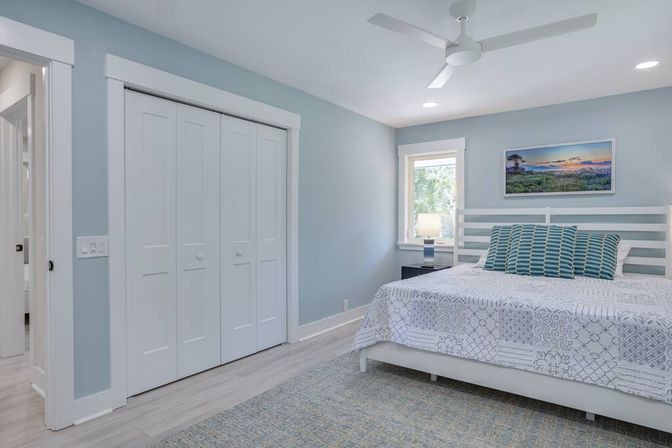 Light, airy coastal-style bedroom with pale blue walls, white slatted bed and patterned quilt, teal striped pillows, white ceiling fan, bedside lamp by a sunny window, double white closet doors and landscape artwork