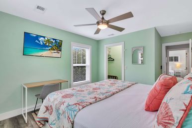 Beach-themed bedroom with mint-green walls, seashell quilt and coral pillows, ceiling fan, wall-mounted TV, small desk and window — cozy coastal guest room