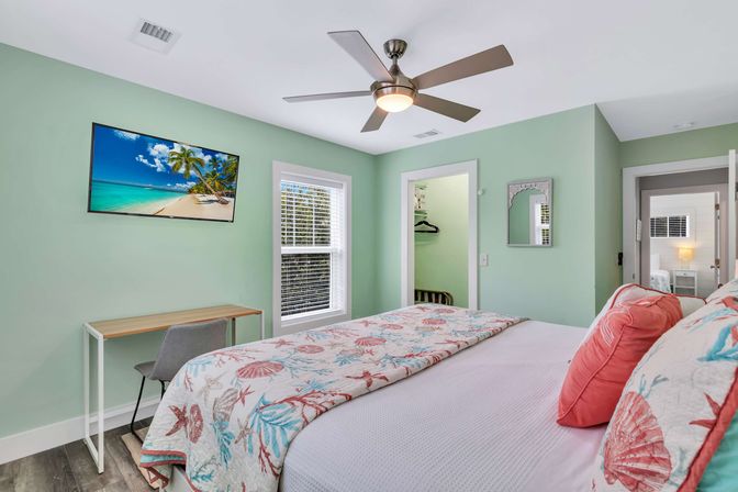 Beach-themed bedroom with mint-green walls, seashell quilt and coral pillows, ceiling fan, wall-mounted TV, small desk and window — cozy coastal guest room