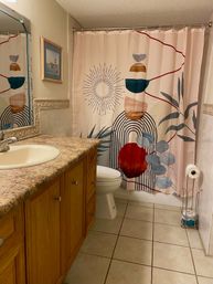 Cozy home bathroom with oak vanity and speckled countertop, tiled floor, white toilet, framed art and a vibrant abstract shower curtain.