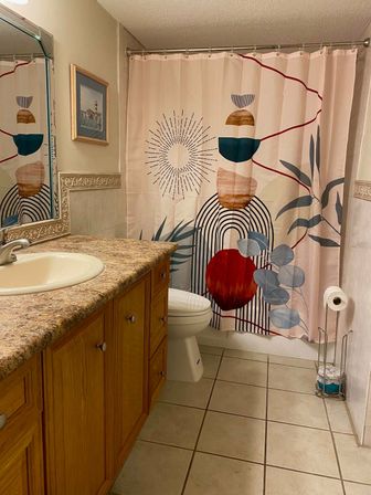 Cozy home bathroom with oak vanity and speckled countertop, tiled floor, white toilet, framed art and a vibrant abstract shower curtain.