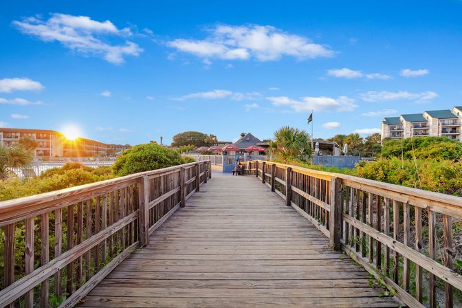 Sunlit wooden boardwalk through coastal dunes and palm trees leading to seaside restaurants and oceanfront hotels at sunset under a bright blue sky.