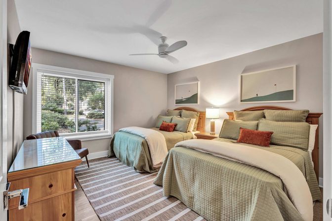 Cozy twin bedroom in a residential home with two beds dressed in sage-green quilted bedding, rust accent pillows, striped rug, wooden nightstand and lamp, ceiling fan, large window showing a leafy street, and minimalist framed art.