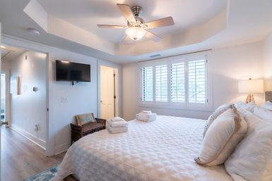 Bright coastal-style bedroom with a king bed in white quilted bedding, neatly folded towels on the foot of the bed, plantation shutters, ceiling fan, wall-mounted TV and a wicker accent chair.