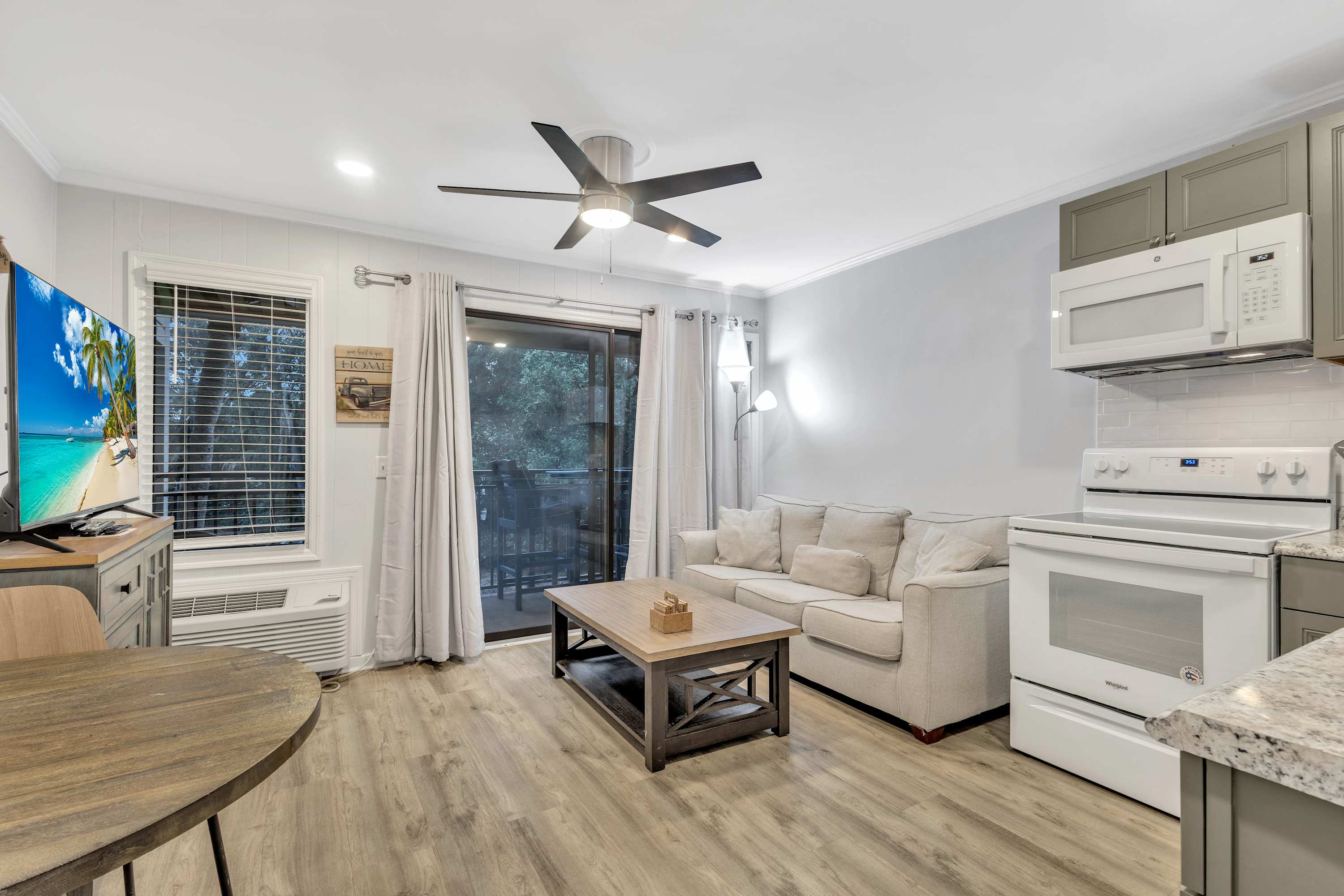 Cozy open-concept apartment living area with beige sofa, wooden coffee table, modern ceiling fan, kitchenette with white stove and microwave, sliding glass door to a balcony, wall-mounted TV and wood-look flooring.