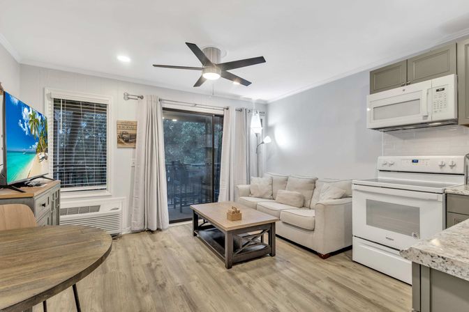 Cozy open-concept apartment living area with beige sofa, wooden coffee table, modern ceiling fan, kitchenette with white stove and microwave, sliding glass door to a balcony, wall-mounted TV and wood-look flooring.