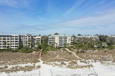 Aerial view of beachfront condominium complex with low-rise buildings, palm trees and wooden boardwalks crossing grassy dunes to a white sandy beach under a bright blue sky.