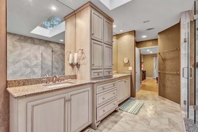 Sunlit luxury master bathroom with double-sink granite vanity, light wood cabinets and central storage tower, skylight, glass-enclosed shower, and marble tile floor in warm neutral tones