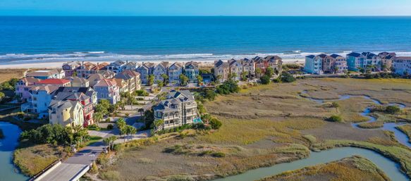 Aerial view of sunlit pastel beachfront homes along a sandy shore, backed by tidal marshes with winding creeks flowing to the blue ocean horizon.
