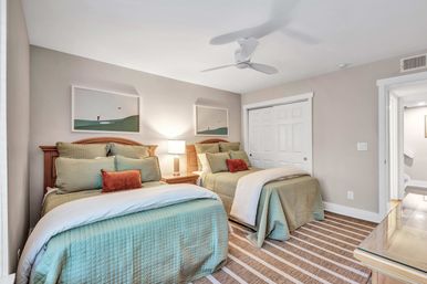 Light, airy guest bedroom with two twin beds dressed in green quilted bedding, rust accent pillows, wooden headboards, central nightstand with lamp, white ceiling fan, sliding closet doors and striped rug.