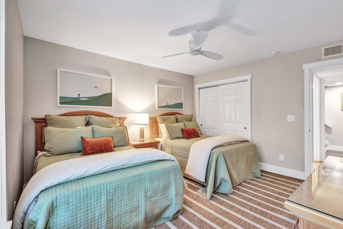 Light, airy guest bedroom with two twin beds dressed in green quilted bedding, rust accent pillows, wooden headboards, central nightstand with lamp, white ceiling fan, sliding closet doors and striped rug.