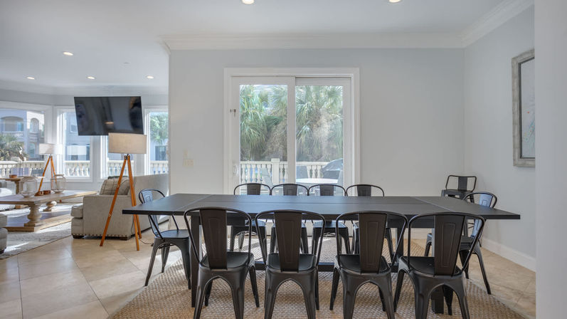 Bright coastal dining room with a long black table and metal industrial chairs for ten, sliding glass doors framing palm trees and a balcony, open to a light-filled living area.
