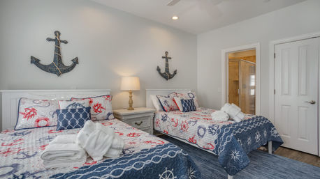 Cheerful coastal-themed bedroom with two twin beds in navy and red nautical quilts, rope-anchor wall decor above a shared nightstand and lamp, folded towels on the beds and an open door to an en-suite bathroom.