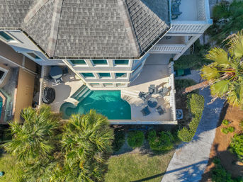 Drone aerial view of a coastal-style three-story home with a turquoise backyard pool, sunlit patio with lounge chairs and surrounding palm trees.